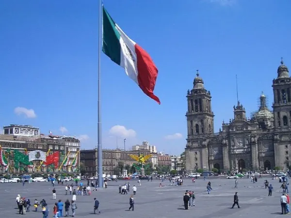Mexico City's historic centre — crowded colonial streets, a cathedral dome rising above the rooftops, street vendors and pedestrians crossing a wide stone plaza under a pale blue sky.