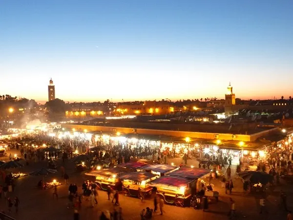 Open-air street market in Mirleft, southern Morocco. Stalls line the dusty road, vendors and shoppers weaving between colourful goods, the warm late-afternoon light catching the dust.