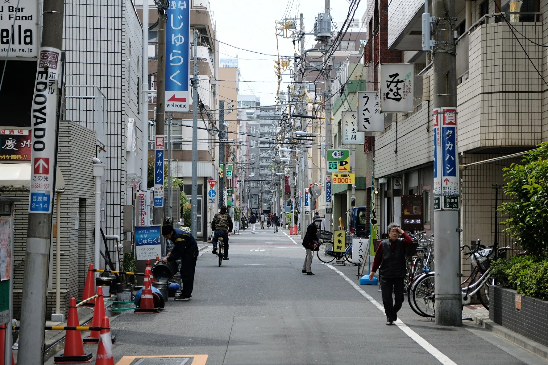 A narrow side street in Nishi-Asakusa, Tokyo. Mid-morning, tall vertical signs in blue and red reaching up toward the tangle of wires overhead. Footsteps on the asphalt, unhurried. A cyclist coasts past, the soft tick of a freewheel. Behind, the metallic ring of a paint can set down on the ground, two voices too far off to make out. The hum of the city sits a few blocks away — traffic, a distant train — but here Tokyo has thinned to a murmur. A crow calls once from a rooftop. The wind moves through the wires. The neighborhood goes about its quiet morning, and the city holds it gently in place.