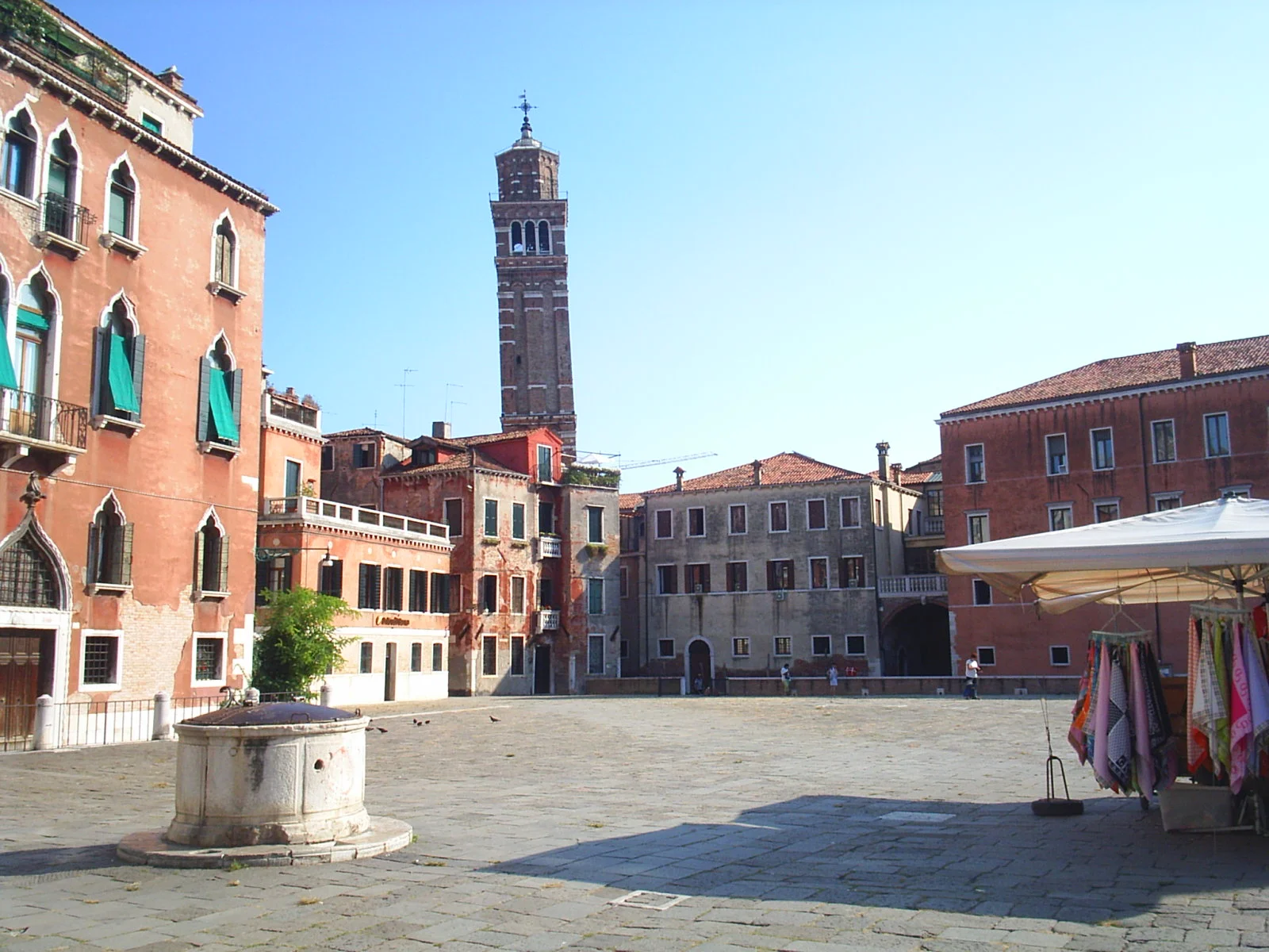 A sunlit piazza in Venice, Italy. Stone paving, people crossing in every direction, the ornate façades of old buildings surrounding the square on all sides.