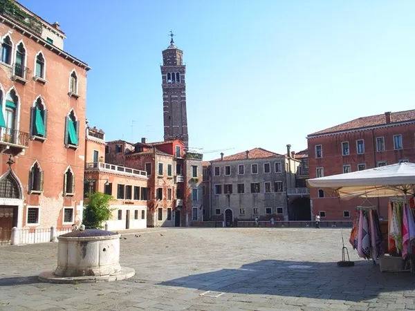 A sunlit piazza in Venice, Italy. Stone paving, people crossing in every direction, the ornate façades of old buildings surrounding the square on all sides.